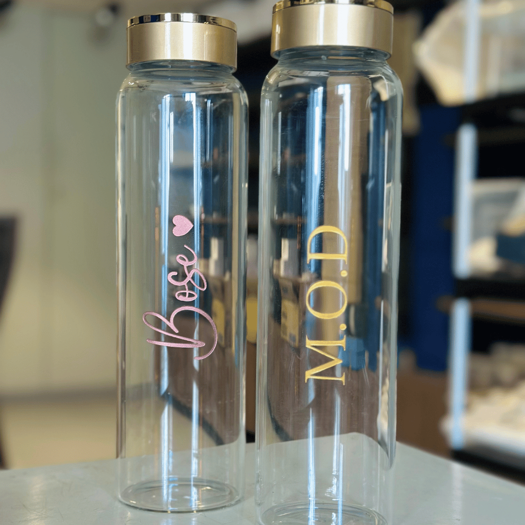 Two clear water bottles with gold and pink text on a blurred indoor background