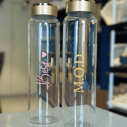 Two clear water bottles with gold and pink text on a blurred indoor background