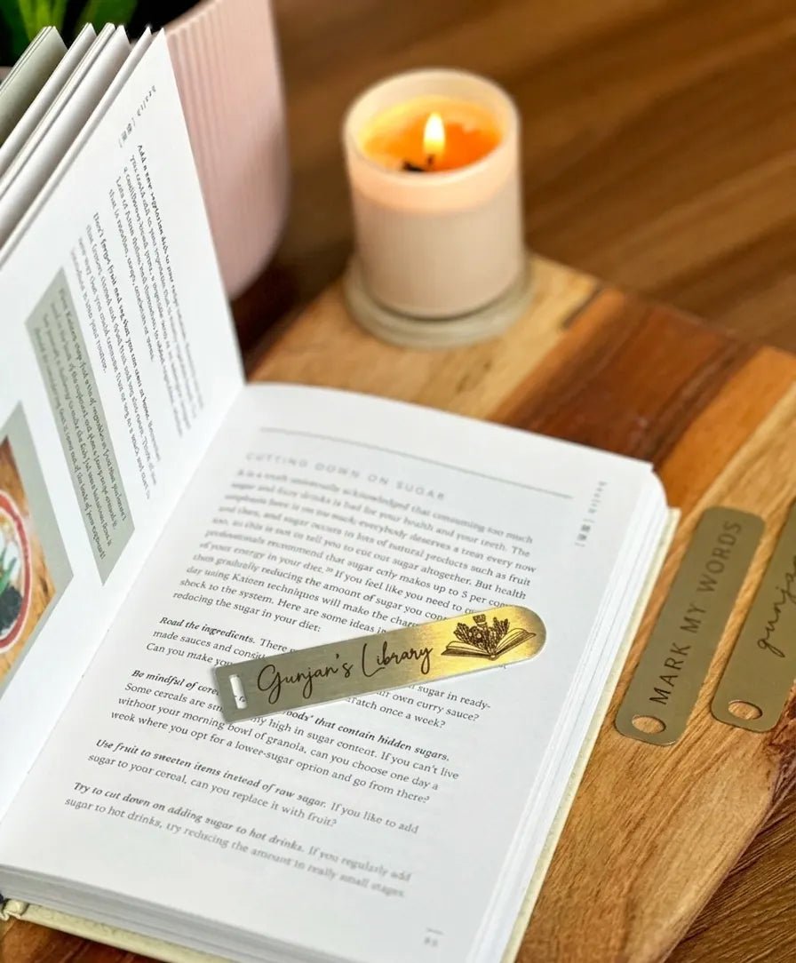 A close-up shot of an engraved gold-toned metal page marker strip resting across the pages of a novel, styled with aesthetic warm lighting.