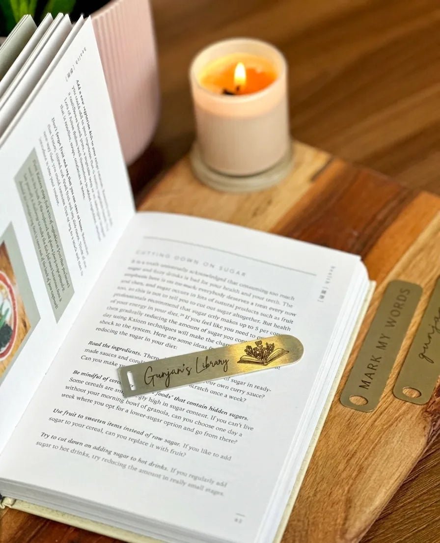 An open book resting on a wooden tray with a custom brass metal page marker showcasing an engraved script name