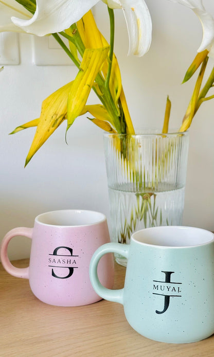 Two ceramic coffee mugs, one pink and one blue, with printed initials on them, placed on a table 